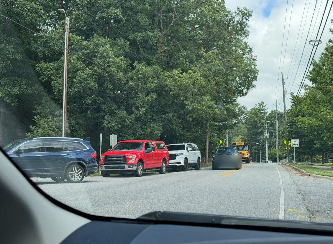Oncoming vehicle straddling the center line on Overlook Road while a queue of stopped cars waits to turn into the school, with a school bus behind them