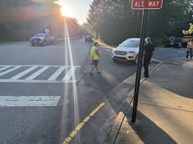 An Asheville Police officer stands at the Pinchot Drive and Red Fox Circle all-way stop during morning arrival while the crossing guard escorts a student, August 2026